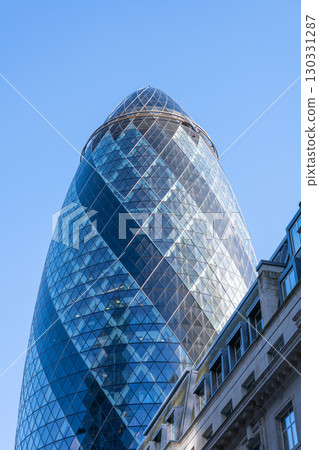 The Gherkin, a modern skyscraper in London's financial district, showcases its unique architectural design against a clear blue sky. Its glass exterior reflects the surrounding cityscape. 130331287