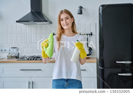 Smiling young woman in gloves with rag cleaning spray looking at camera in kitchen 130332005