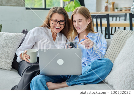 Middle aged mother and daughter 18,19 years old sitting together at home looking at laptop Middle aged mother and daughter 18,19 years old sitting together at home looking at laptop 130332022