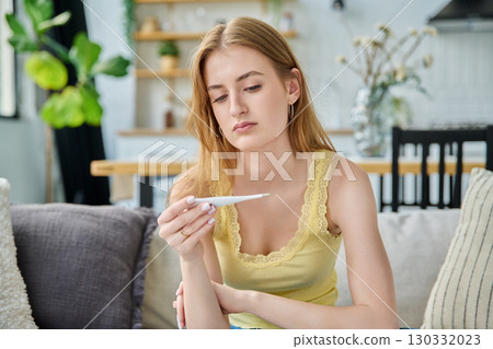 Sad upset teenage girl looking at thermometer sitting at home Sad upset teenage girl looking at thermometer sitting at home 130332023