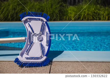 Manual pool vacuum head with blue bristles placed on the edge of a clean swimming pool, designed for scrubbing and suction during routine maintenance. 130332622