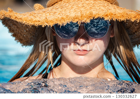 Smiling teenage girl cooling off in a bubbling pool, wearing reflective sunglasses and a wide straw hat under the bright summer sun Smiling teenage girl cooling off in a bubbling pool, wearing reflective sunglasses and a wide straw hat under the bright summer sun 130332627