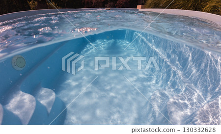 Underwater and above view of a clear blue swimming pool with curved steps and sunlight reflections on the floor and walls, evoking calm and leisure. 130332628