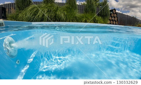 Powerful water jet bubbles underwater in a backyard swimming pool, surrounded by lush greenery and modern wooden fence. 130332629