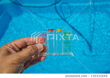 A person holds a clear pool water tester with pH and chlorine indicators above a sparkling blue swimming pool during regular maintenance on a sunny day. A person holds a clear pool water tester with pH and chlorine indicators above a sparkling blue swimming pool during regular maintenance on a sunny day. 130332649