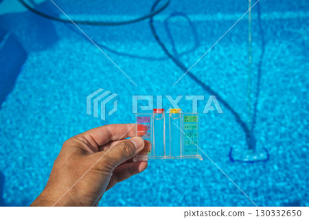 Hand holding a pool water test kit with pH and chlorine scale above a clear blue swimming pool, showing tools used for pool maintenance and water quality control. 130332650