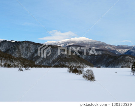 Lake Akimoto in Urabandai in winter. The lake surface is frozen white, and on the opposite shore, the snow-capped Mount Azuma shines white, creating an impressive and beautiful scene against the blue sky. 130333529