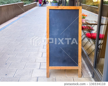 Wooden chalkboard sign standing on a pedestrian street next to cafe terrace and pink chairs. Urban commerce, communication and business display symbolizing customer service and advertising copy space. Wooden chalkboard sign standing on a pedestrian street next to cafe terrace and pink chairs. Urban commerce, communication and business display symbolizing customer service and advertising copy space. 130334110
