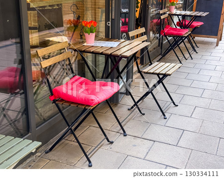 Cafe terrace with wooden tables, chairs and bright pink cushions near potted flowers. Hospitality, lifestyle and dining experience symbolizing service, customer comfort and urban leisure. 130334111