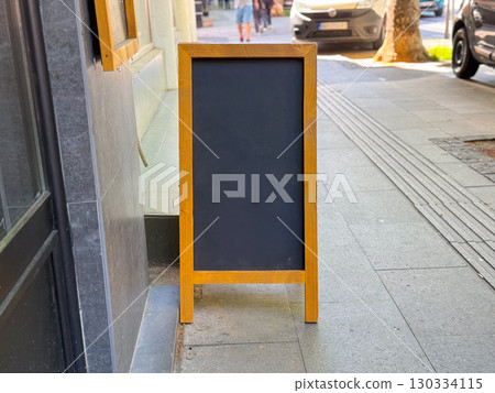 Blank chalkboard sign standing outside on a sidewalk near a cafe entrance, copy space. Urban street, business and advertisement display representing communication, information and customer promotion. Blank chalkboard sign standing outside on a sidewalk near a cafe entrance, copy space. Urban street, business and advertisement display representing communication, information and customer promotion. 130334115