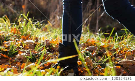 Low view to female feet in boots going along trail on fallen dry leaves. Legs of young woman stepping along path outdoor. Girl walking at nature on sunny autumn day. Slow motion 130334181