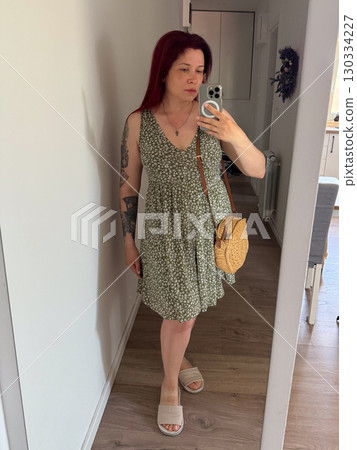 A woman in a green dress with a floral pattern takes a photo of herself in mirror. Summer look, emphasis on lightness and femininity. Concept of fashion, style, self-presentation A woman in a green dress with a floral pattern takes a photo of herself in mirror. Summer look, emphasis on lightness and femininity. Concept of fashion, style, self-presentation 130334227