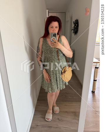 A woman in a green dress with a floral pattern takes a photo of herself in mirror. Summer look, emphasis on lightness and femininity. Concept of fashion, style, self-presentation 130334228