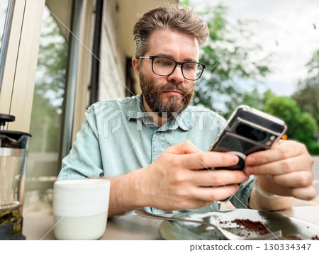 Man using smartphone at outdoor cafe table after eating cake. Modern lifestyle moment of technology, leisure and communication in urban dining environment. Man using smartphone at outdoor cafe table after eating cake. Modern lifestyle moment of technology, leisure and communication in urban dining environment. 130334347