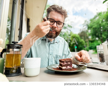 Man eating cake at outdoor cafe with tea pot and cup. Lifestyle and leisure dining experience of everyday relaxation and authentic food enjoyment. Man eating cake at outdoor cafe with tea pot and cup. Lifestyle and leisure dining experience of everyday relaxation and authentic food enjoyment. 130334348