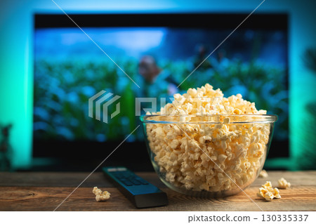 A glass bowl of popcorn and remote control in the background the TV works. Evening cozy watching a movie or TV series at home 130335337