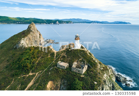 Lighthouse stands on rocky coastline, overlooking the ocean with mountains in the background during a clear day 130335598