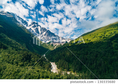Majestic snow-capped Caucasus mountains rise above lush green valleys and a winding river on a sunny day in the wilderness 130335680