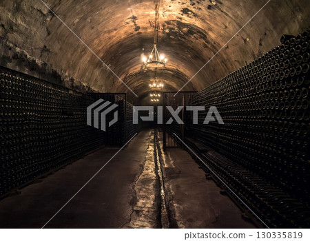 Dark underground wine cellar featuring endless rows of bottles illuminated by soft chandeliers Dark underground wine cellar featuring endless rows of bottles illuminated by soft chandeliers 130335819