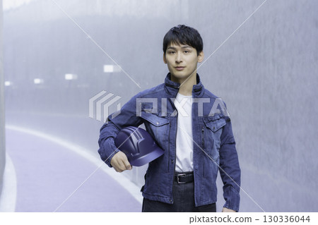 Portrait of a young man wearing denim work clothes and holding a helmet 130336044