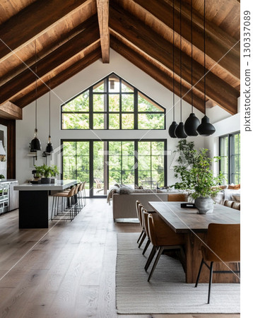 A stunning modern farmhouse interior with a vaulted wooden ceiling and large windows overlooking lush greenery. The open-plan space features a kitchen island, dining table, and living area. 130337089