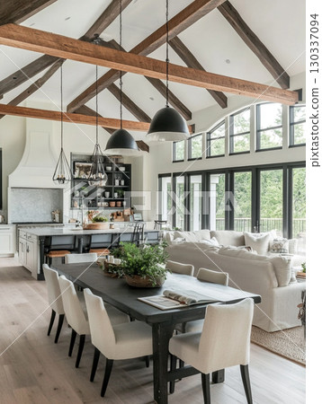 A stunning modern farmhouse interior with a vaulted wooden ceiling and large windows overlooking lush greenery. The open-plan space features a kitchen island, dining table, and living area. 130337094