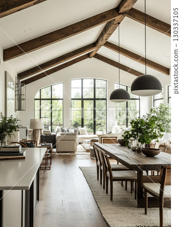 A stunning modern farmhouse interior with a vaulted wooden ceiling and large windows overlooking lush greenery. The open-plan space features a kitchen island, dining table, and living area. 130337105