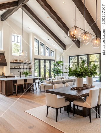 A stunning modern farmhouse interior with a vaulted wooden ceiling and large windows overlooking lush greenery. The open-plan space features a kitchen island, dining table, and living area. 130337107