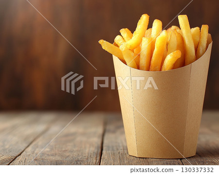 A cup of crispy, golden french fries stands upright on a rustic wooden table. The focus is on the delicious, fast-food snack against a dark, blurred background. 130337332