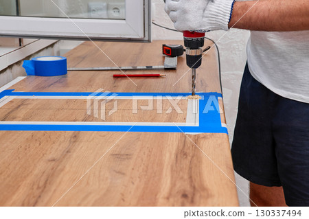 repairman using electric drill for cutting hole for the sink in the kitchen countertops, installation new modern kitchen 130337494