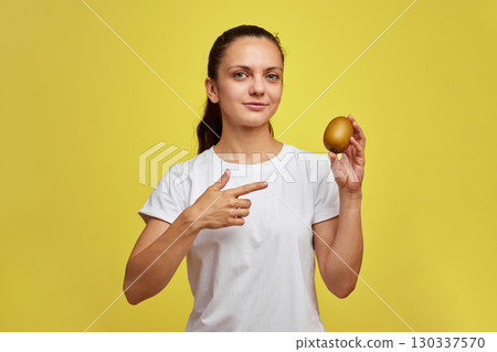 Happy woman in white t-shirt holding kiwi on studio yellow background 130337570
