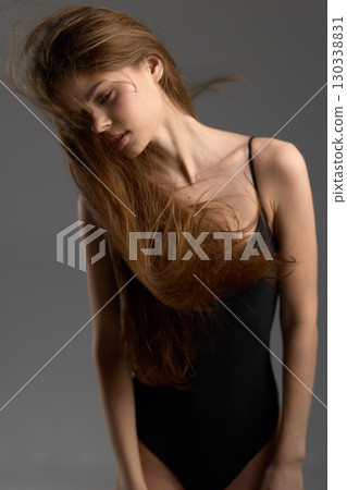 Young woman with long flowing hair in black swimsuit posing against neutral gray background with soft lighting. 130338831