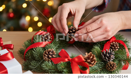 Christmas wreath being decorated by hand with pinecones and red ribbons, festive crafting scene with warm lights in the background 130340144