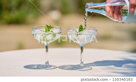 Two refreshing summer cocktails with ice and mint in coupe glasses, with sparkling water being poured on sunny outdoor table. 130340163