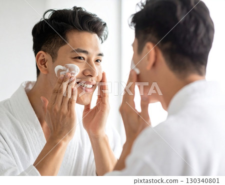 Smiling man applying face cleanser foam in front of bathroom mirror, skincare routine. High quality photo 130340801
