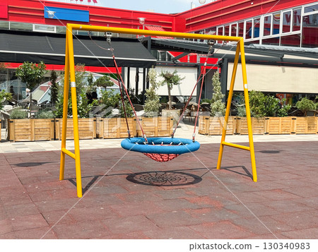 Yellow swing frame with round blue net seat on playground. Childhood leisure, recreation, and outdoor play equipment in urban public space. 130340983