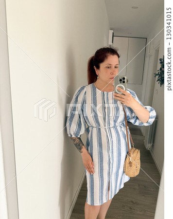 A woman in a striped dress takes a photo of herself in mirror. Summer look, emphasis on lightness and femininity. Concept of fashion, style, self-presentation 130341015