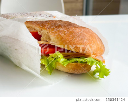 Fresh sandwich with lettuce, tomato, cheese, and ham wrapped in paper on table. Nutrition, lifestyle, and fast food expressed through diet, snack, and consumer culture. Fresh sandwich with lettuce, tomato, cheese, and ham wrapped in paper on table. Nutrition, lifestyle, and fast food expressed through diet, snack, and consumer culture. 130341424