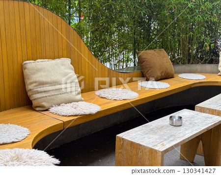 Wooden curved bench with pillows and small table placed in outdoor terrace. Hospitality, leisure, and lifestyle expressed through decor, design, and natural greenery Wooden curved bench with pillows and small table placed in outdoor terrace. Hospitality, leisure, and lifestyle expressed through decor, design, and natural greenery 130341427