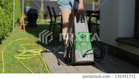 Woman pulling a high-pressure washer in the backyard of a house. Woman pulling a high-pressure washer in the backyard of a house. 130341751