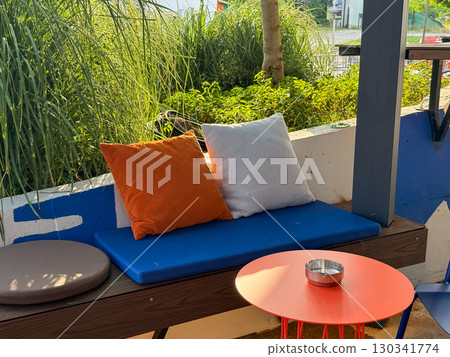 Outdoor cafe corner with green plants, pillows, and small red table with ashtray. Leisure, comfort, and hospitality connected with urban lifestyle and relaxation culture. 130341774