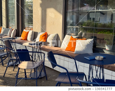 Modern cafe terrace with orange and white pillows on benches near blue tables. Comfort, leisure, and social interaction through contemporary hospitality and outdoor culture. 130341777