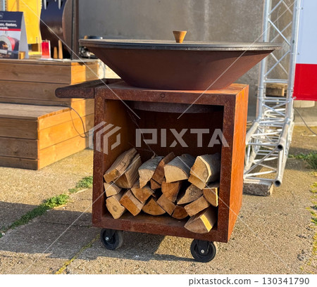 Outdoor grill with firewood stored below standing on wheels in sunlight. Culinary culture, preparation, and lifestyle linked with barbecue, cooking, and outdoor events. Outdoor grill with firewood stored below standing on wheels in sunlight. Culinary culture, preparation, and lifestyle linked with barbecue, cooking, and outdoor events. 130341790