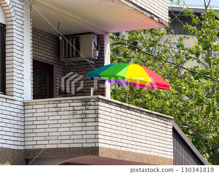 Rainbow umbrella standing open on a sunny balcony with brick walls and green trees. Urban lifestyle, leisure, and everyday culture in city housing architecture. 130341819