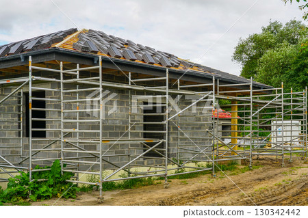 Scaffolding around unfinished brick house with roof tile installation in residential construction site 130342442