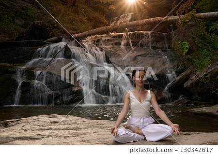 Young woman sitting in lotus pose under tropical waterfall meditating with calm strength and mindfulness fully connected to nature energy tranquility and inner balance Young woman sitting in lotus pose under tropical waterfall meditating with calm strength and mindfulness fully connected to nature energy tranquility and inner balance 130342617