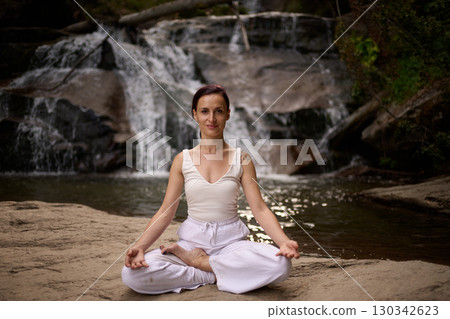 Young woman sitting in lotus pose under tropical waterfall meditating with calm strength and mindfulness fully connected to nature energy tranquility and inner balance 130342623