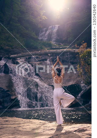 Woman doing yoga tree pose in front of waterfall at sunrise peaceful meditation in nature  130342640