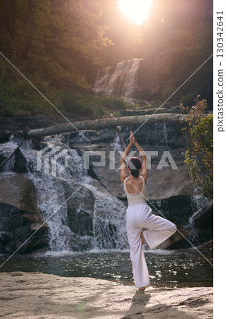 Woman doing yoga tree pose in front of waterfall at sunrise peaceful meditation in nature Woman doing yoga tree pose in front of waterfall at sunrise peaceful meditation in nature 130342641