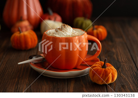 Cozy pumpkin latte in a pumpkin-shaped mug with whipped cream and spices, surrounded by autumn decor. Cozy pumpkin latte in a pumpkin-shaped mug with whipped cream and spices, surrounded by autumn decor. 130343138